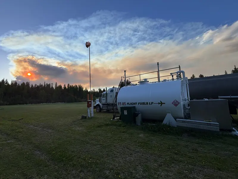 A bulk fuel tank and tanker truck on a grass field under a smoky orange sunset
