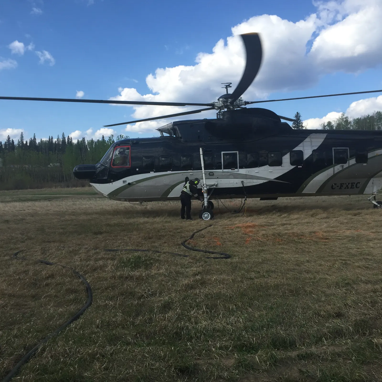 A heavy-lift helicopter being fuelled from a hose laid across a grass field, two crew standing by
